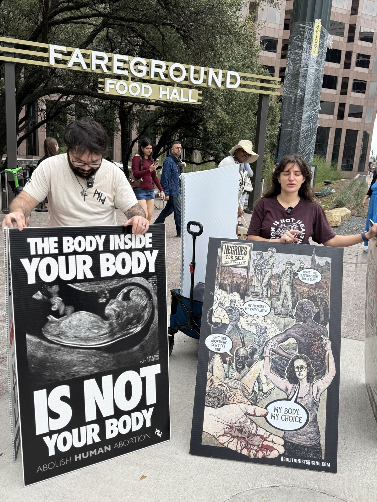 Two people holding large signs under a food hall sign